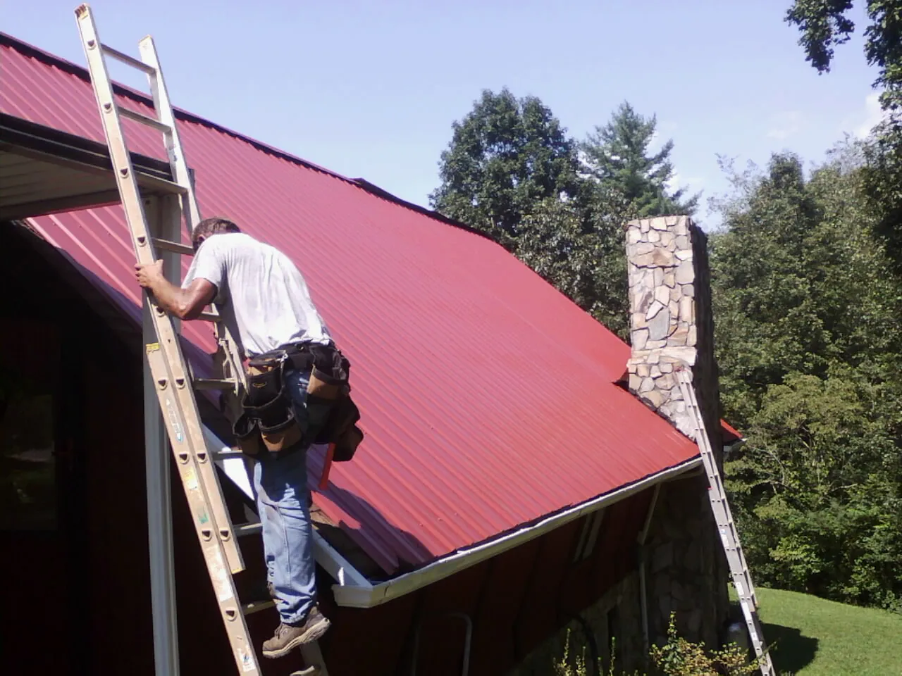 Roofer on a ladder beside a red metal roof and stone chimney — Soffit & Fascia Repair by Roofing Brothers in Port Trevorton
